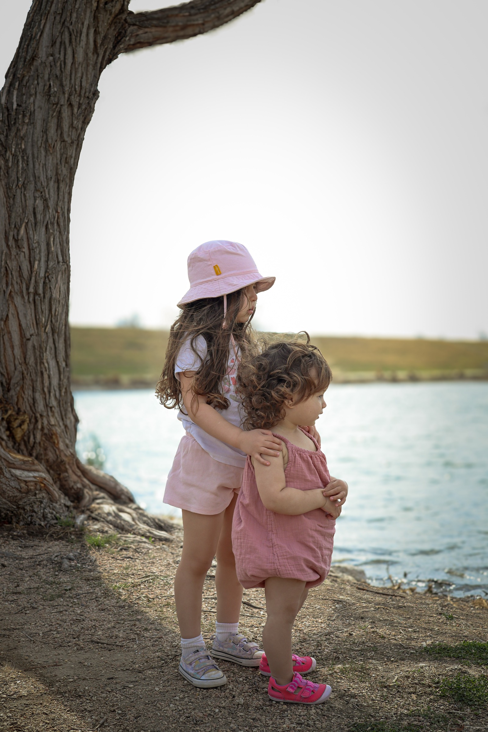 Two little girls standing together by the water, the older sister with her arm around the younger