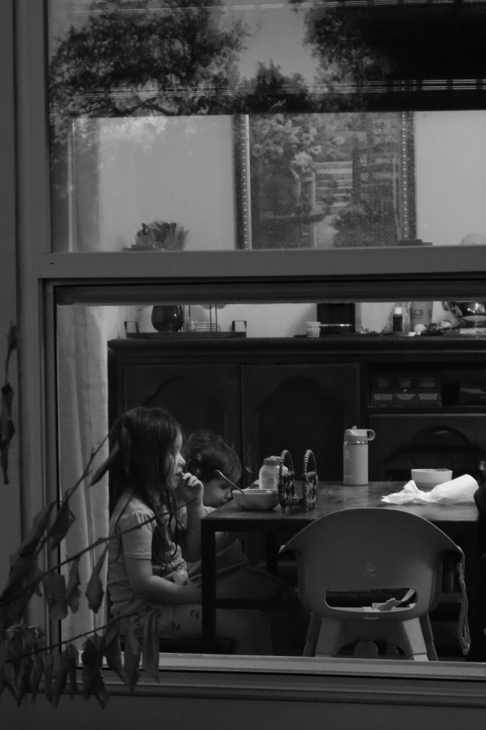 Black and white photograph of two children eating breakfast, seen through a window